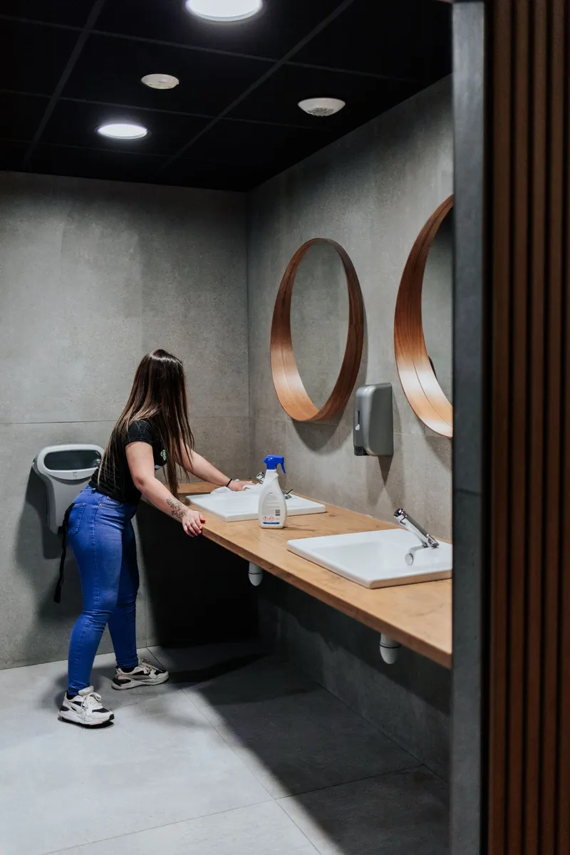 Employée de Pro-Net nettoyant les lavabos dans des toilettes modernes avec des miroirs ronds à Paris.