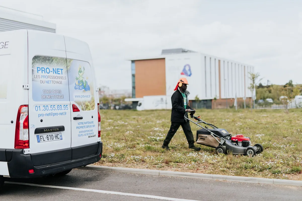 Employé de Pro-Net tondant l'herbe près d'un véhicule de l'entreprise affichant des informations de contact, dans un espace extérieur urbain.